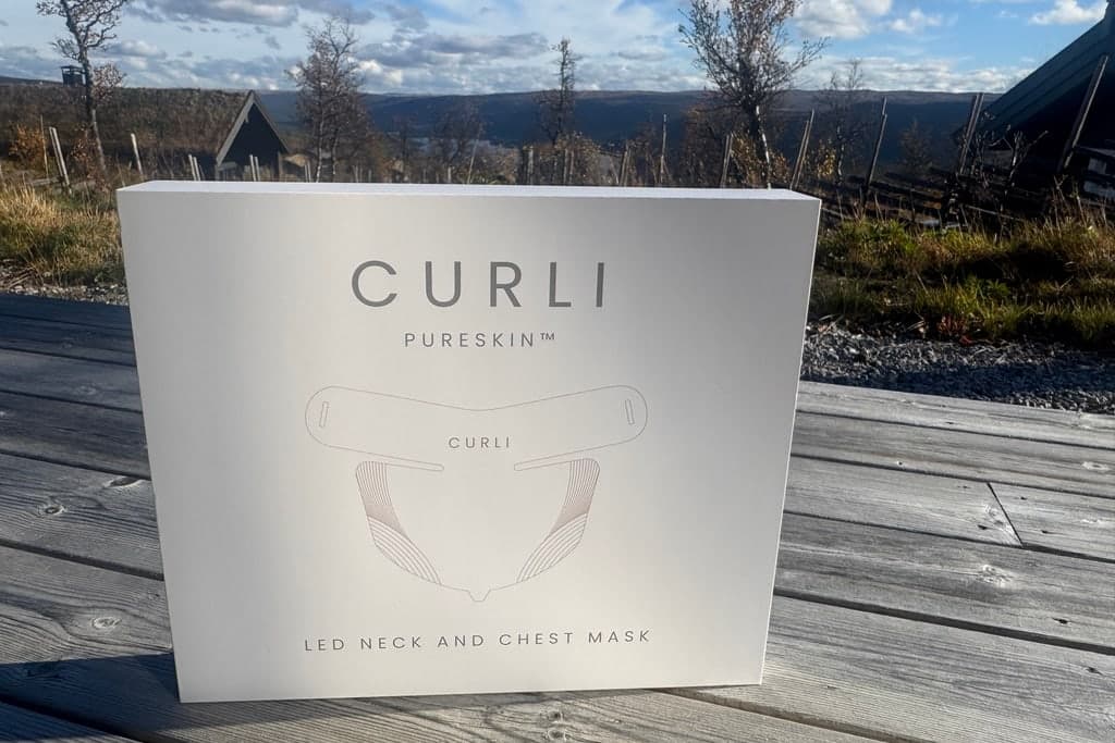 Esken til CURLI PureSkin LED hals- og brystmaske på et trebord utendørs, med fjell og himmel i bakgrunnen