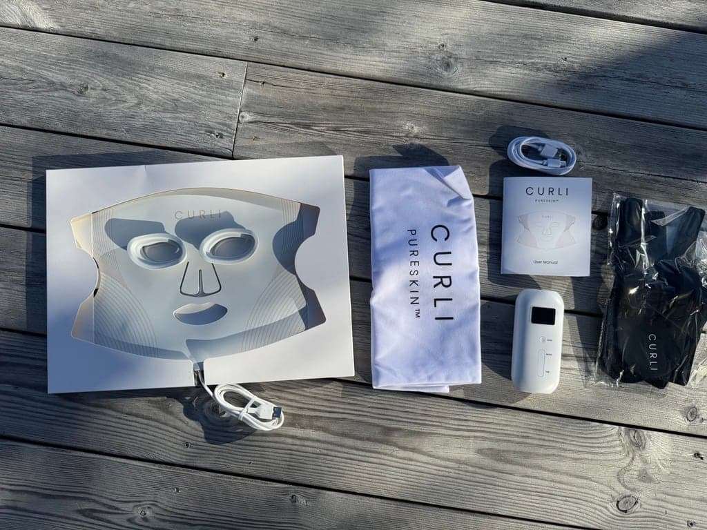 Ansiktsmasken i innlegget fra esken, sammen med oppbevaringspose, USB-kabel, bruksanvisning, kontrollenhet og et svart tilbehør, lagt ut på et trebord